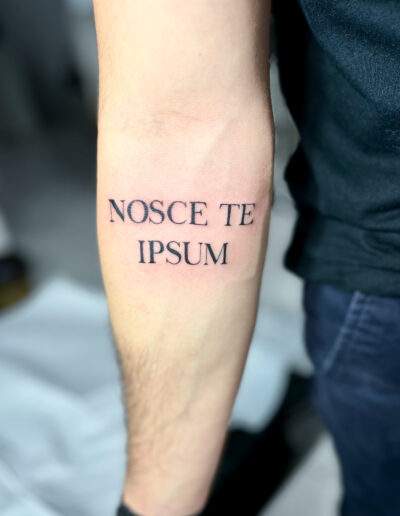 Minimalistyczny, konturowy tatuaż - lettering, napis NOSCE TE IPSUM na przedramieniu wykonany w studiu Omerta Tattoo, Wrocław